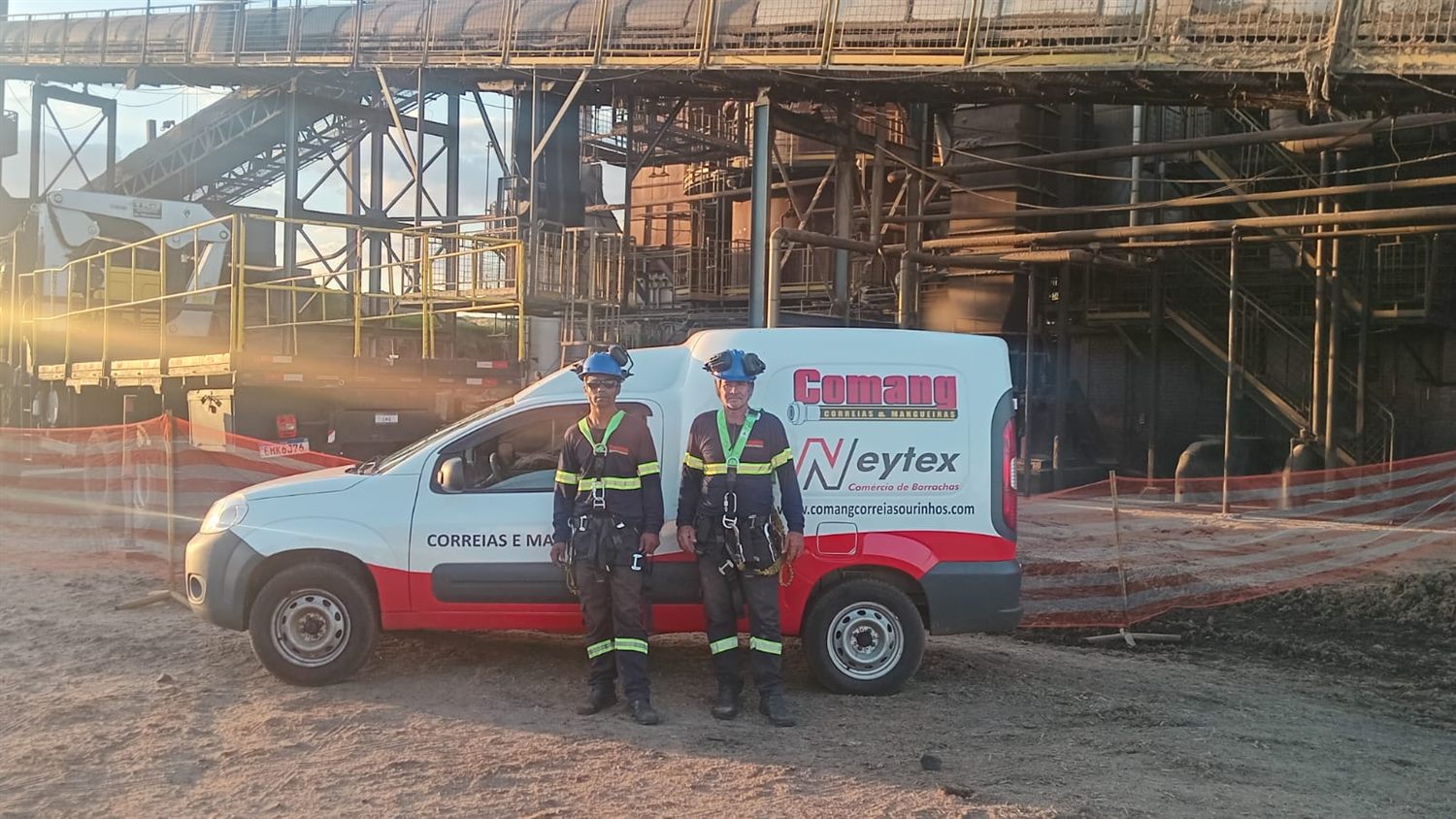 Dois técnicos da Comang de capacete e cinto de segurança em frente ao veículo da empresa, dentro de uma planta industrial